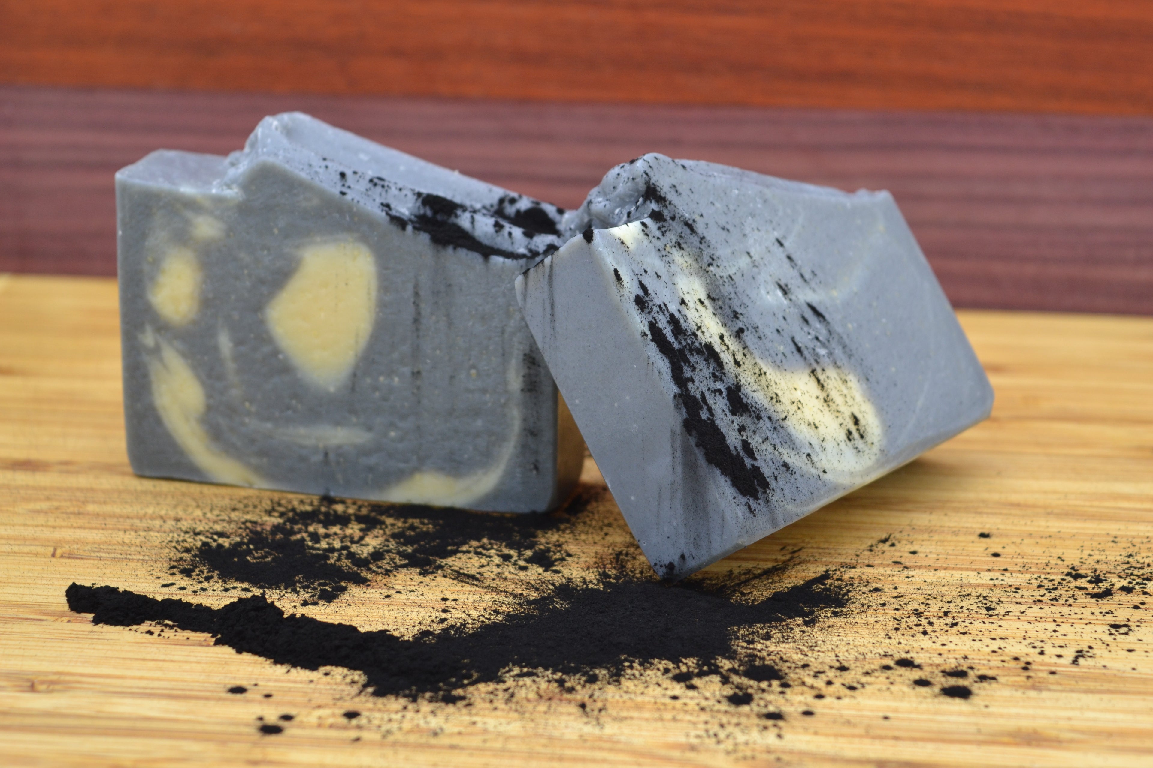 Two pieces of gray soap with black and cream swirls on a wooden surface with charcoal scattered around.